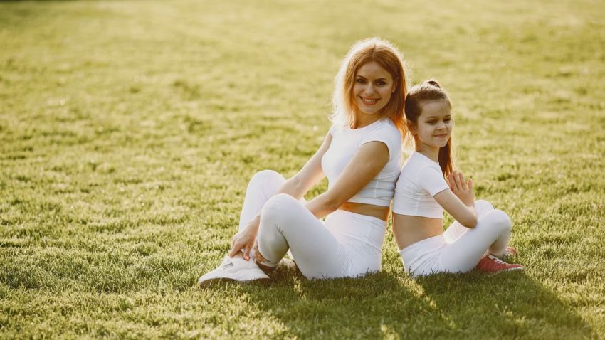 A mother and daughter sit back to back in matching activewear on a sunny grass field, enjoying a summer day together.